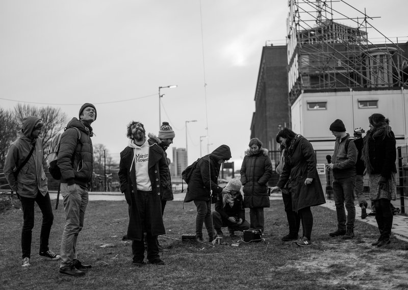 11 people standing in a field; two people looking at the sky, six people looking at connected electronic parts of the 'earth computer'; one person is filming using a handheld video camera and another person holding a microphone; in the middle of the frame