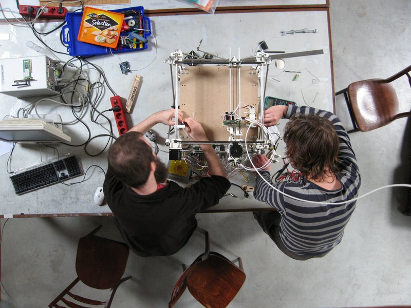 An overhead shot of a big messy table top with a Reprap self replicating 3D printer being constructed by two people, both with their hands inside the half constructed printer; on the left side of the printer is a light grey computer, a monitor, a keyboard
