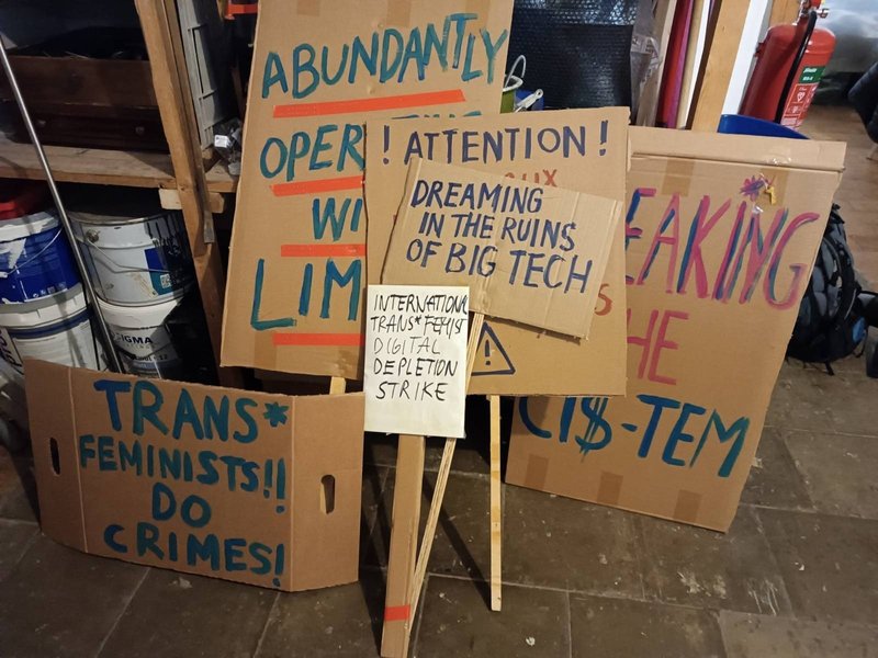 A set of brown five cardboard banners placed on the floor of an interior room, with colourful handwritten messages in capital letters, reading; trans*feminists! Do crimes!; Abundantly operating within limits; International Trans*feminist Digital Depletion